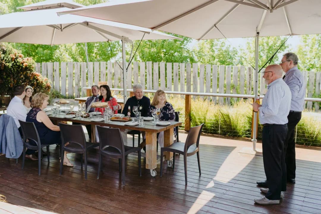 Guests sit at an outdoor reception table beneath umbrellas while a man speaks.