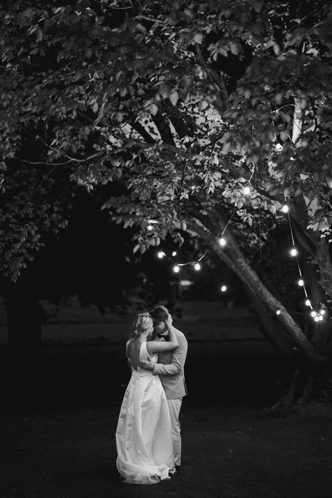 Black and white photograph of the couple embracing beneath a tree decorated with lights.