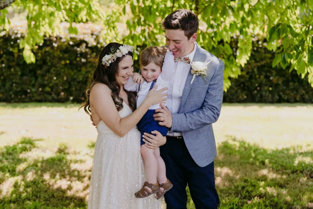 Couple smiles together while holding a young child beneath a leafy tree.