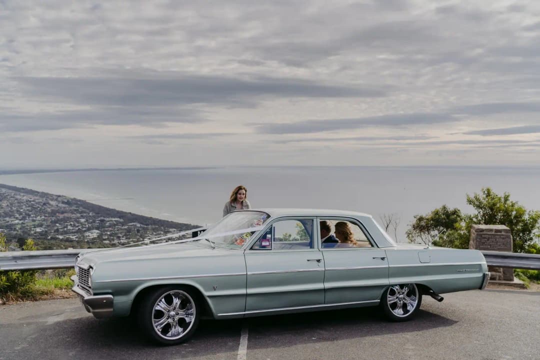 Vintage pale green car parked at a coastal lookout beside the bride.