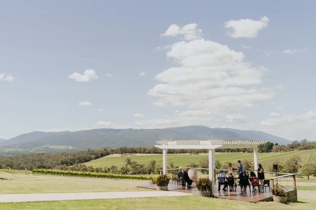Wide view of an outdoor ceremony beneath a white pergola with hills beyond.