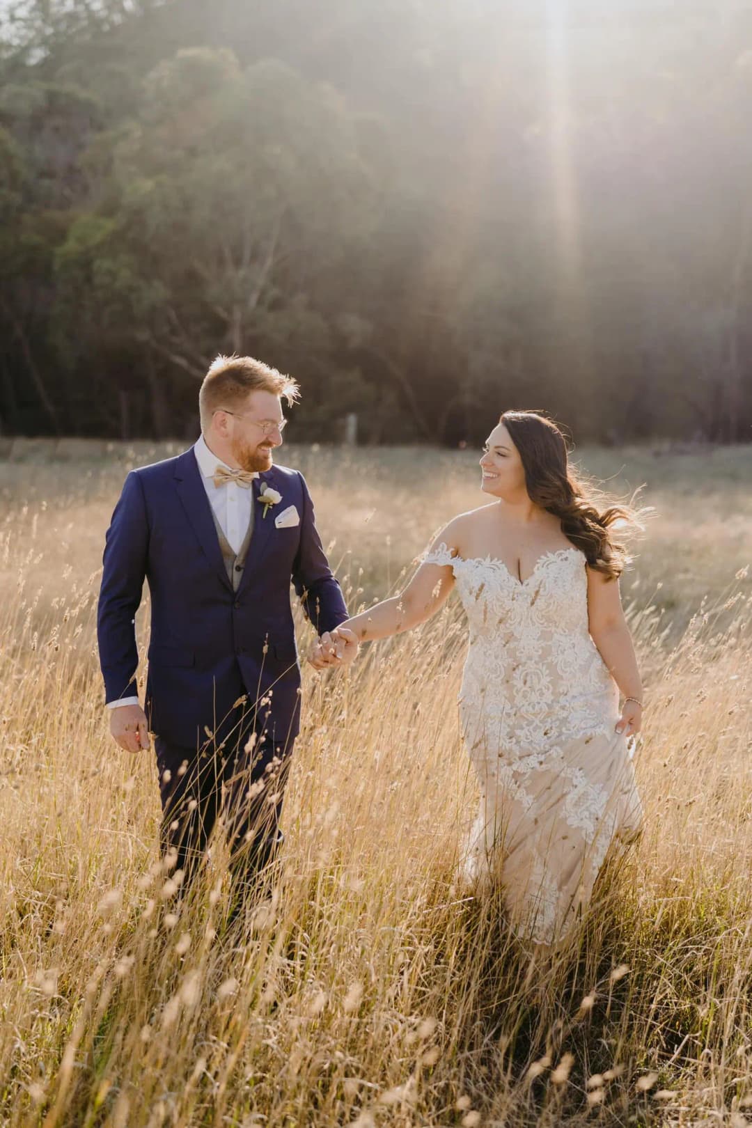 Couple walks hand in hand through tall grass in warm sunlight.