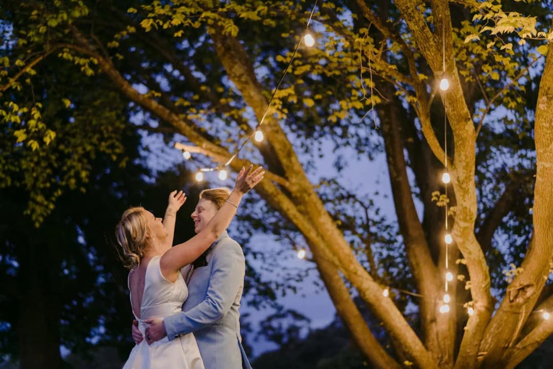 Couple embraces beneath a tree with the bride raising an arm toward the lights.