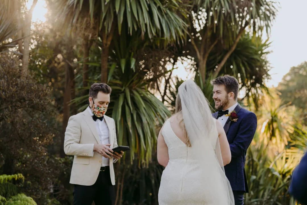 Rear view of the bride facing the groom as a masked celebrant reads outdoors.