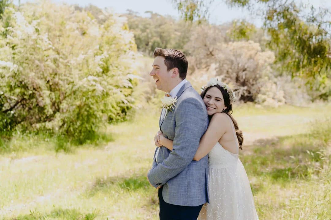 Bride hugs the groom from behind in a sunny garden.