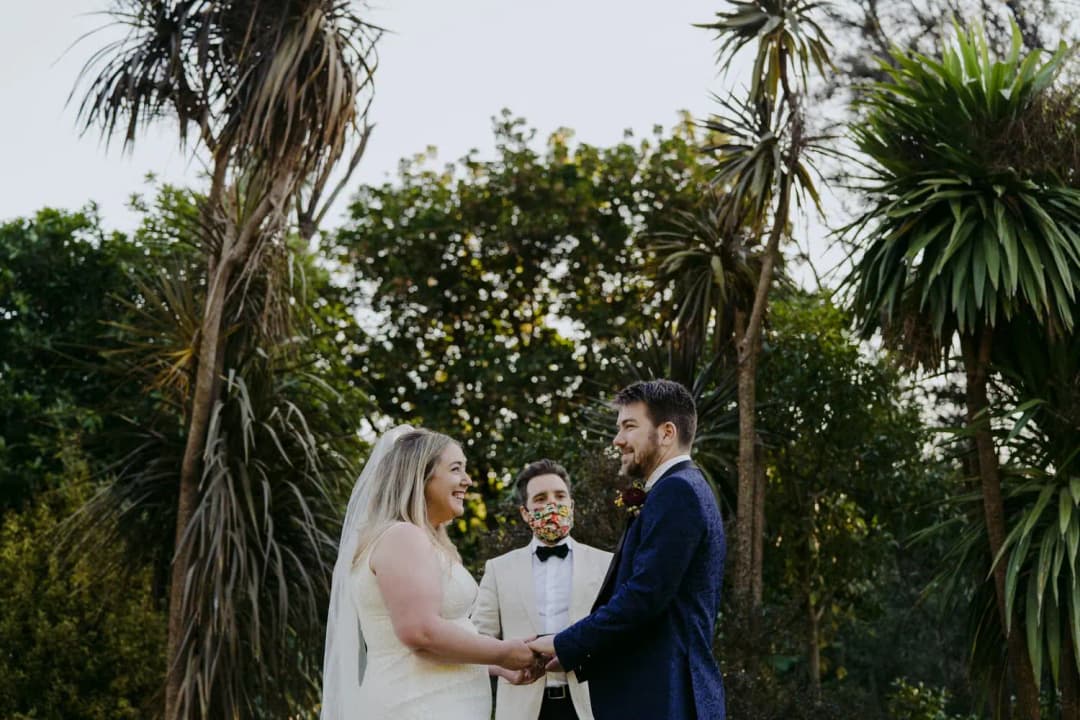 Couple holds hands in a leafy garden beside a masked celebrant.