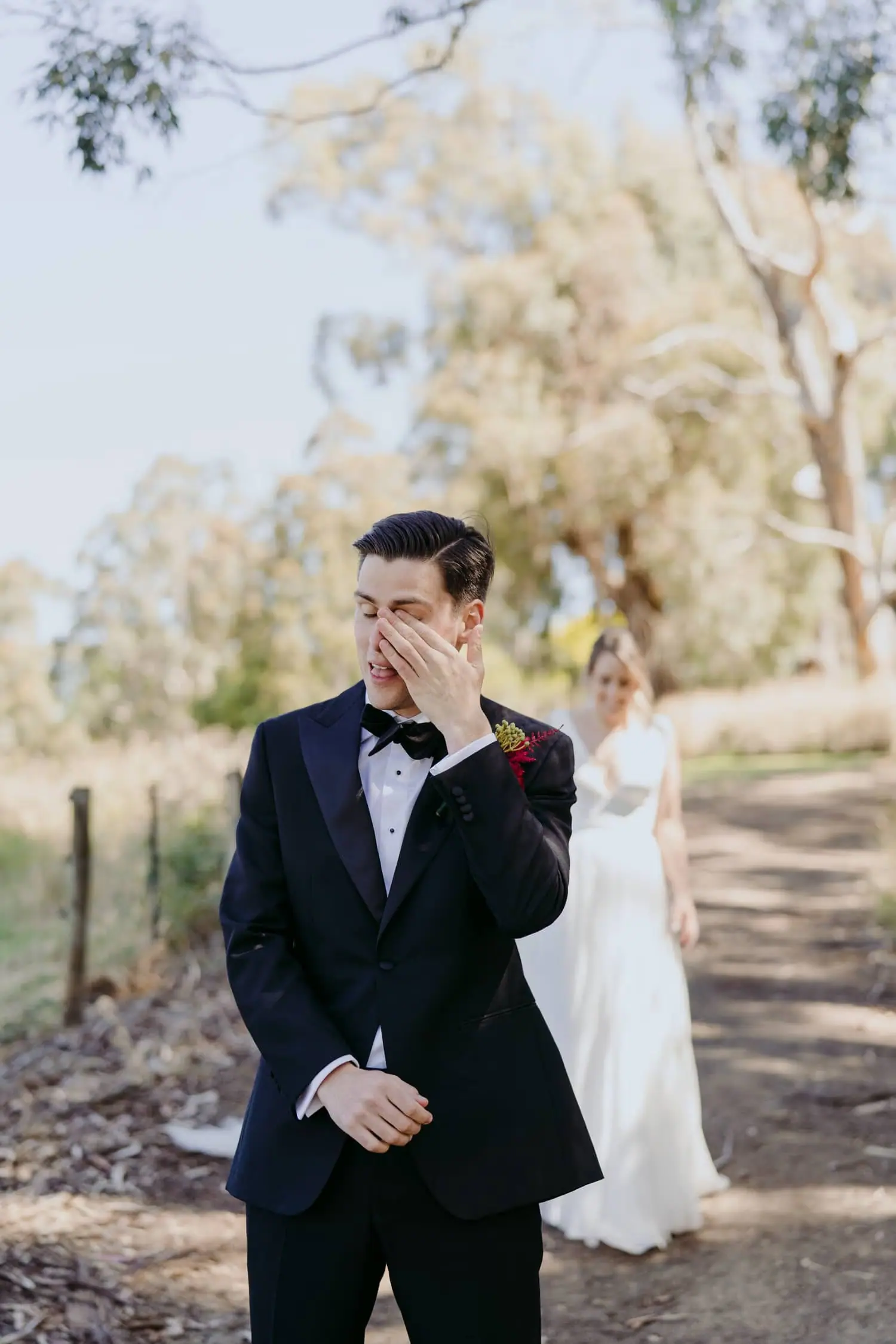 Groom wipes his eye as the bride approaches behind him on a shaded path.