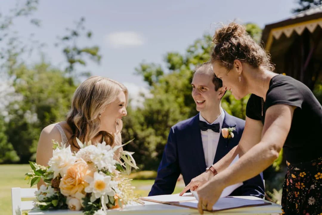 Bride and groom smile as a woman assists them with signing wedding paperwork.