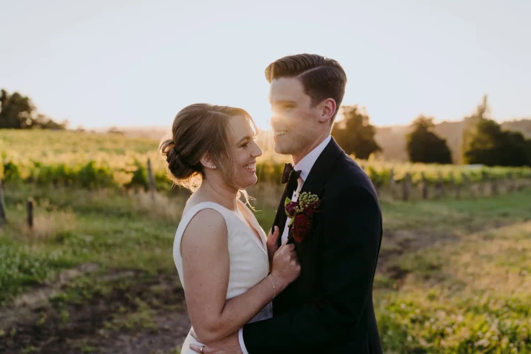 Couple smiles close together beside vineyard rows at sunset.