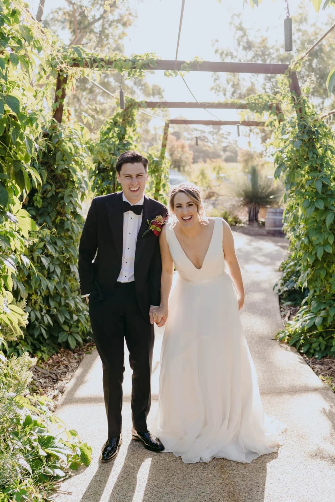 Couple walks along a garden path beneath a vine-covered pergola.