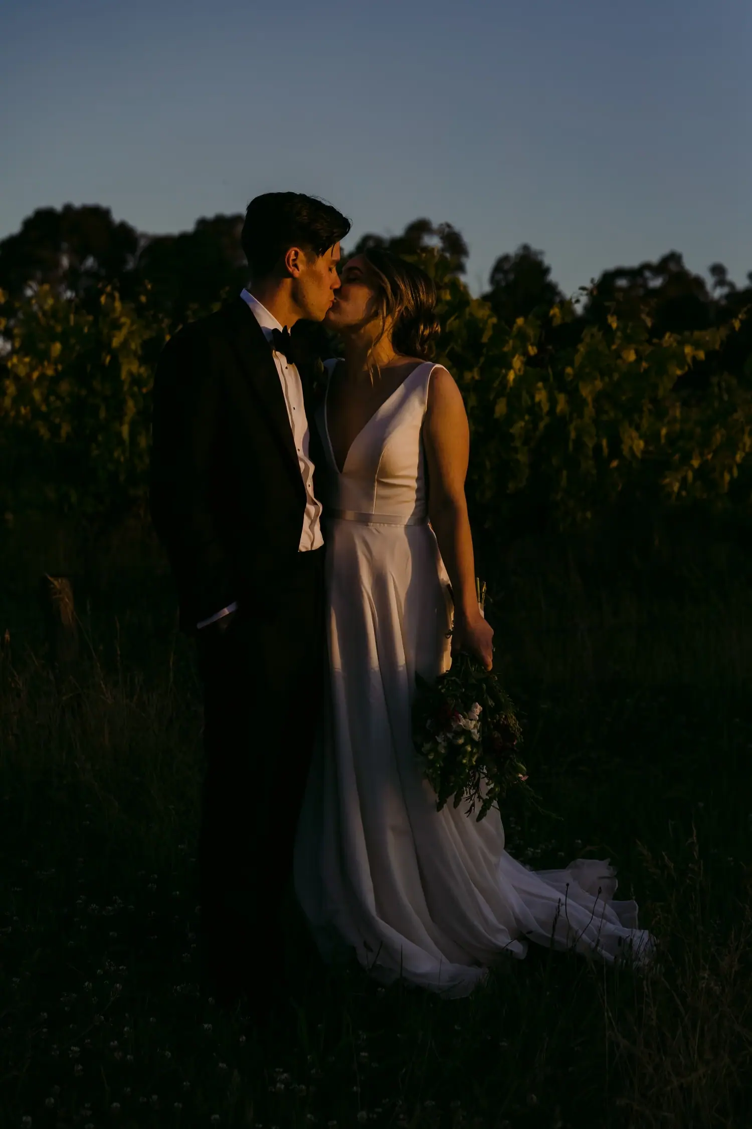 Couple kisses in a field beneath a deep blue evening sky.