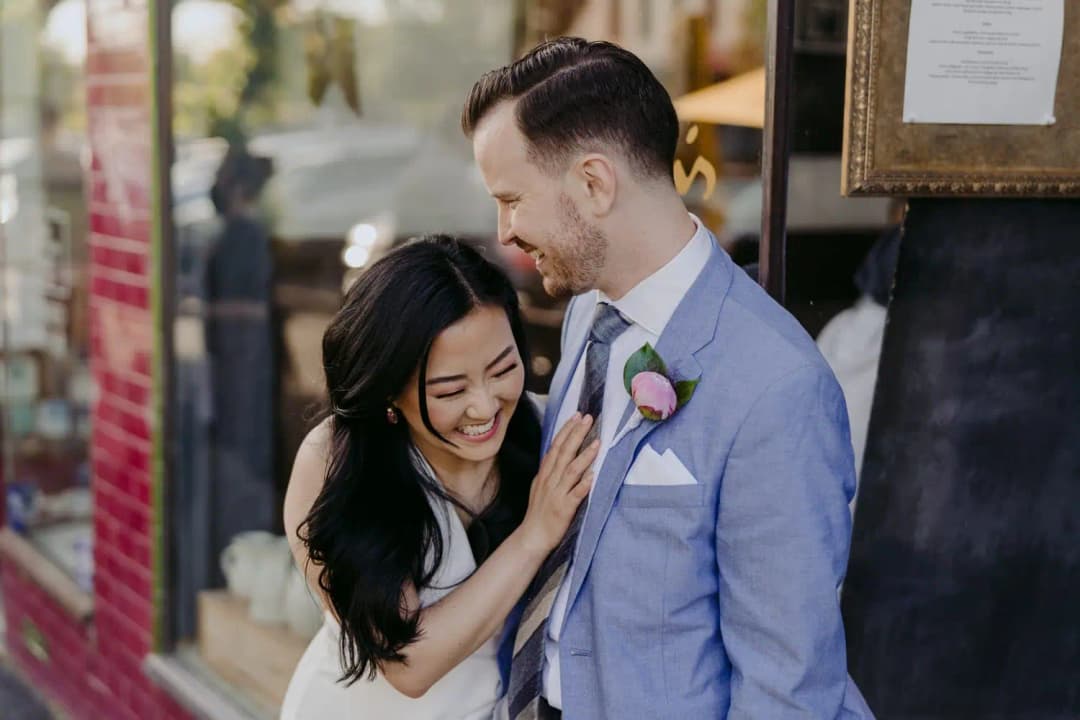 Bride laughs as the groom kisses her forehead outside a building.