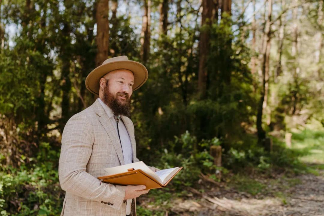 Celebrant in a brimmed hat holds an open book in a wooded setting.