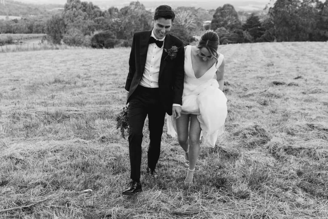 Black and white photograph of the couple walking through grass with the bride gathering her skirt.