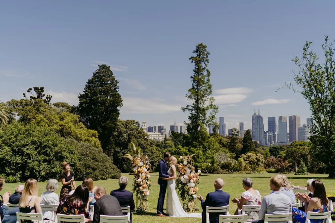 Wide view of a garden wedding ceremony with a city skyline beyond the trees.