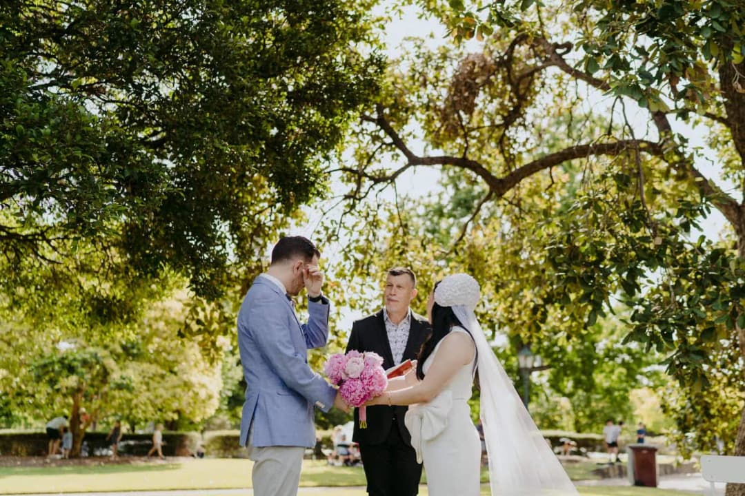 Couple holds hands beneath a leafy tree as the celebrant speaks.