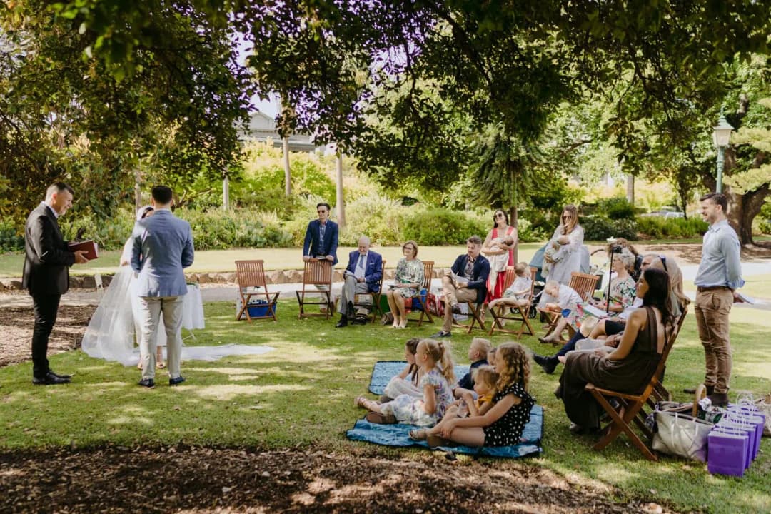 Small wedding ceremony under garden trees with guests on chairs and children seated on the grass.