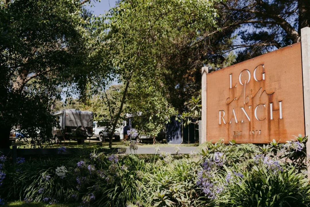Rust-coloured ranch sign beside flowering plants and tall garden trees.