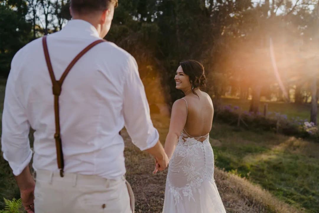 Bride looking back while holding the groom's hand beneath sunlit trees.