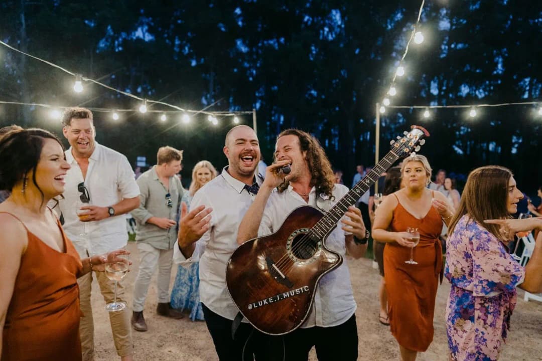 Guitarist singing among guests beneath glowing outdoor string lights.