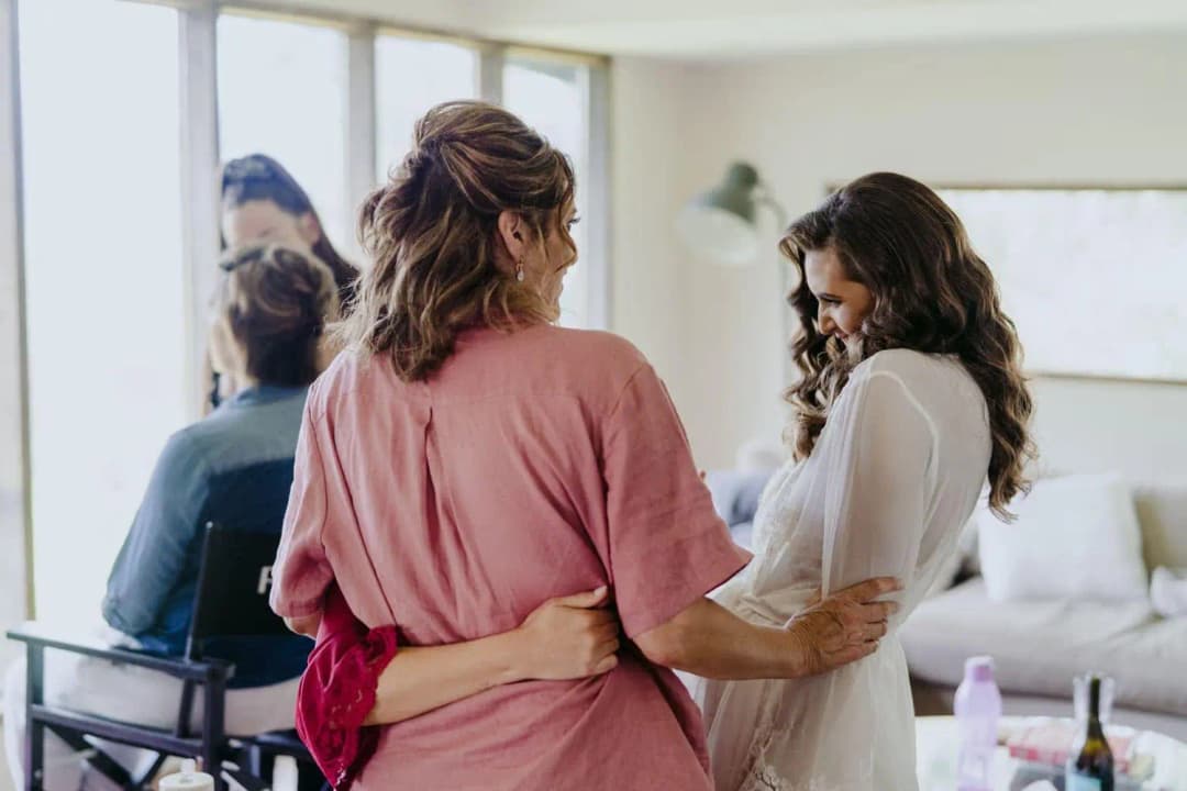 Bride in a white robe standing with a woman in pink while another woman has her hair styled.