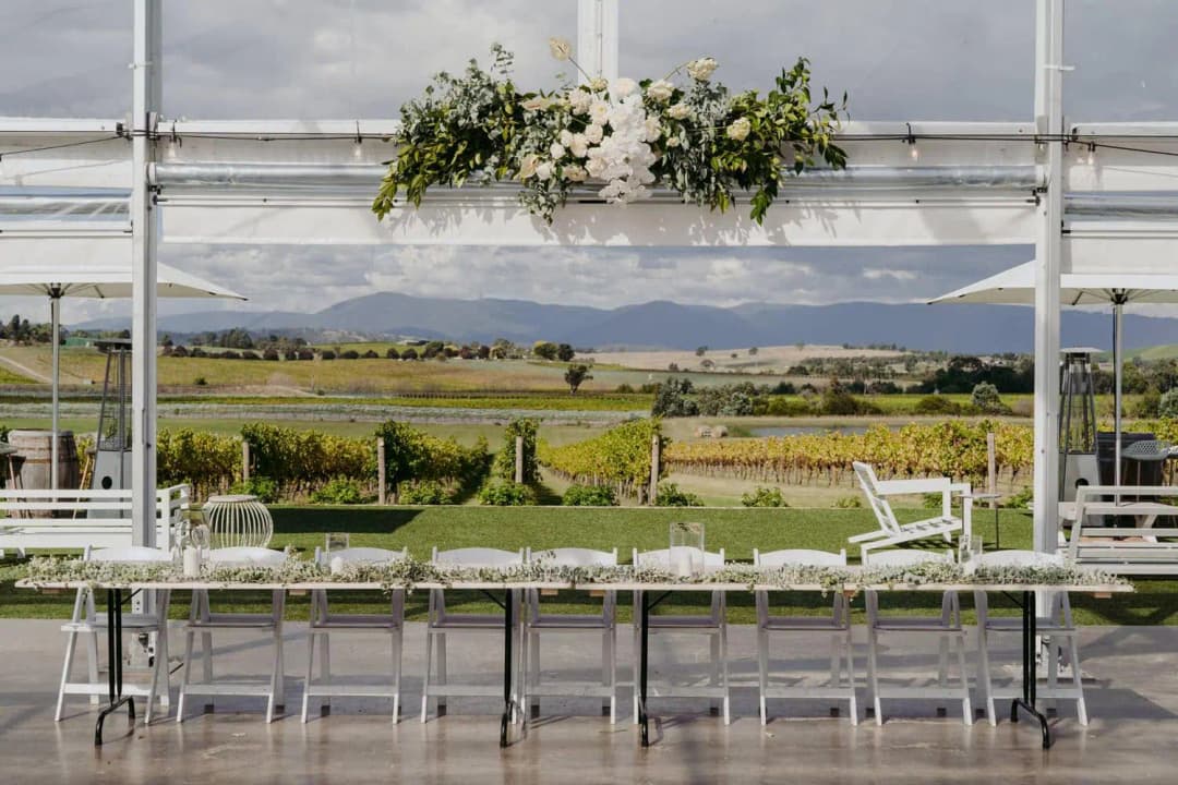 Long reception table beneath hanging flowers in a clear marquee overlooking farmland.