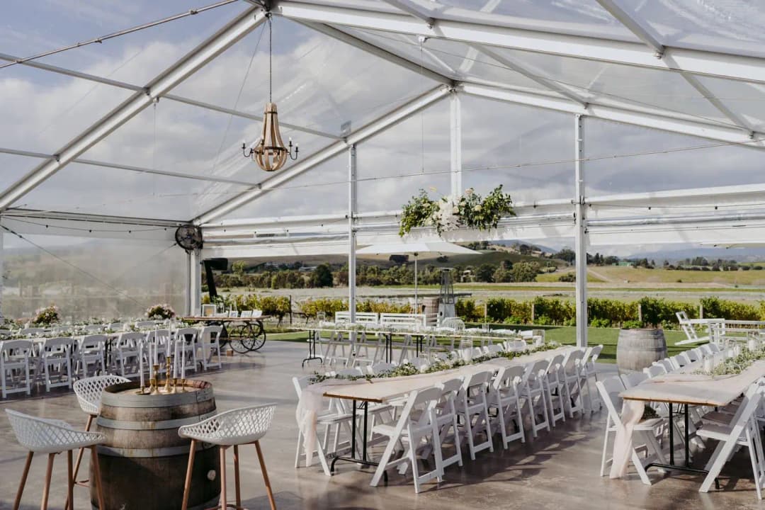 White chairs and reception tables beneath chandeliers in a clear-roof marquee.