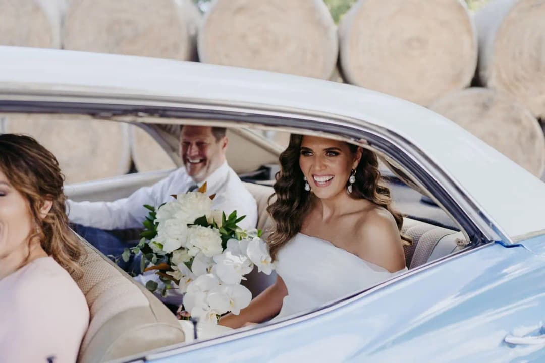 Bride holding her bouquet and smiling from inside a blue vintage car.