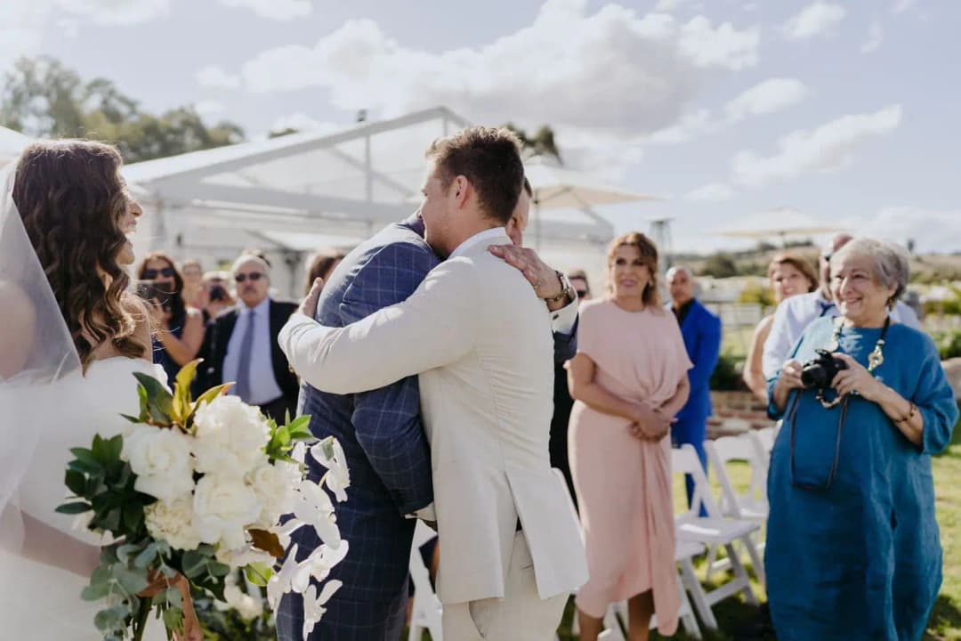 Groom hugging a man in a blue suit while the bride stands nearby.