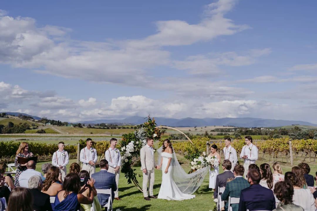 Wide view of the wedding party at a floral arch with seated guests and mountains beyond.