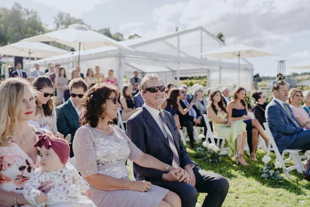 Guests seated on white chairs at the outdoor ceremony, including a woman holding a baby.