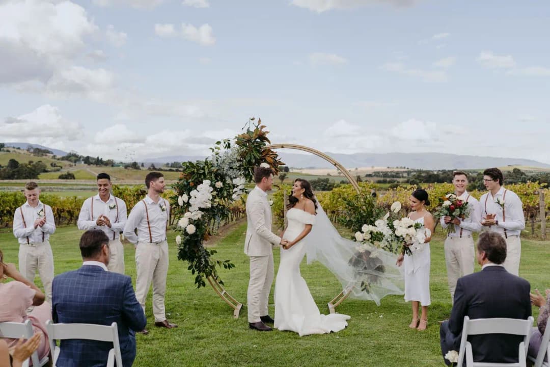Bride and groom facing each other beneath the floral arch while their wedding party watches.