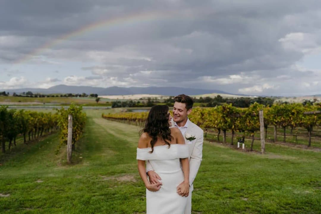 Groom embracing the bride from behind between rows of grapevines.
