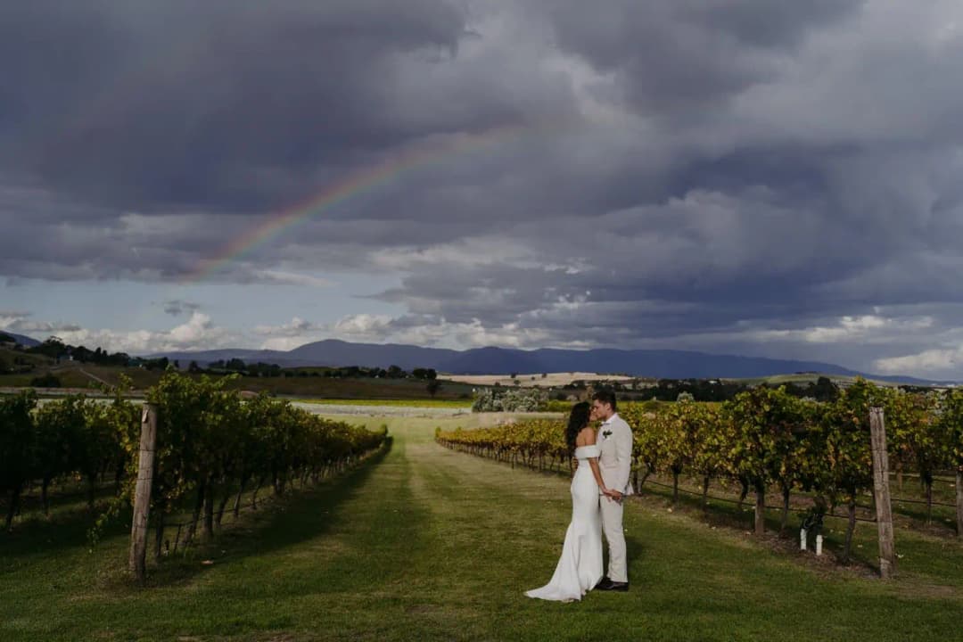 Wide portrait of the couple on a vineyard path beneath dark clouds and a rainbow.