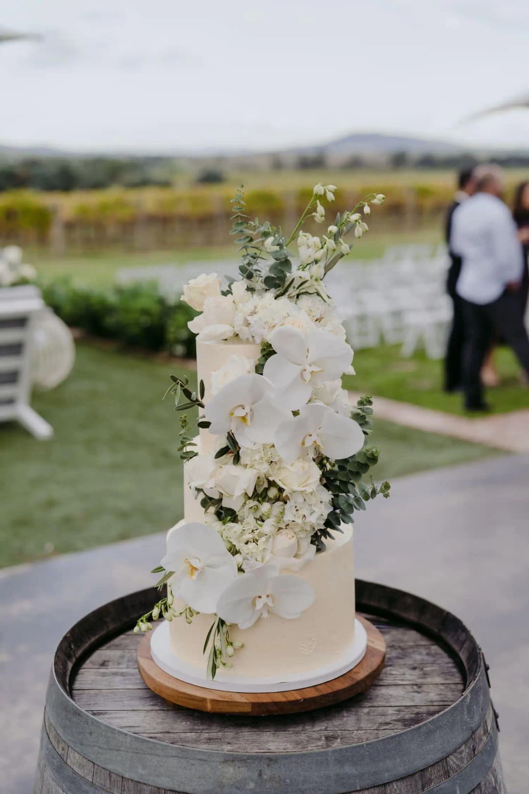 Tall white wedding cake decorated with white flowers on a wooden barrel.