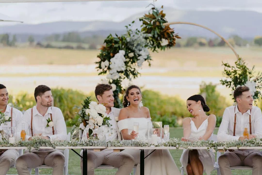 Bride and groom laughing with their wedding party at a greenery-decorated reception table.