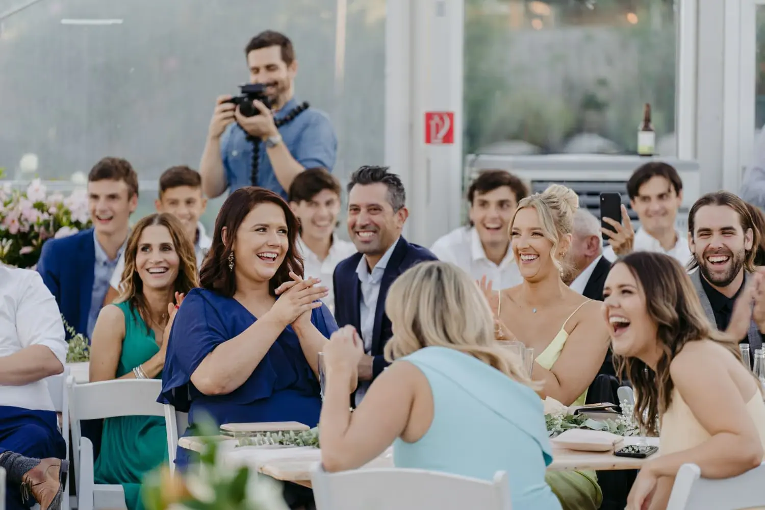 Guests laughing at a reception table while a man behind them photographs the moment.