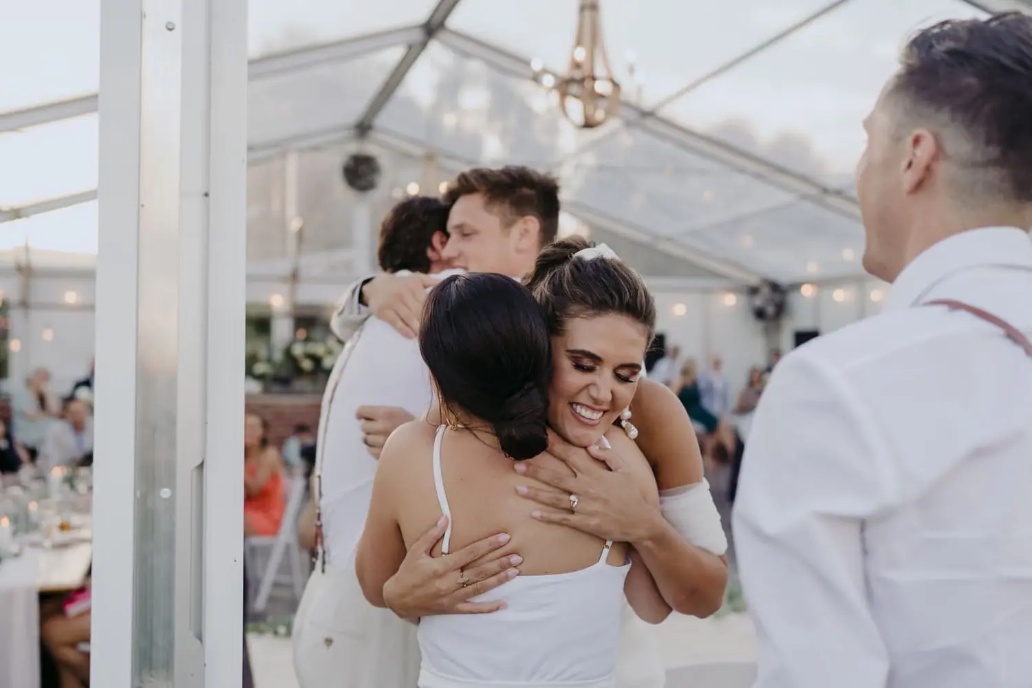 Bride hugging a woman while other wedding party members embrace nearby.