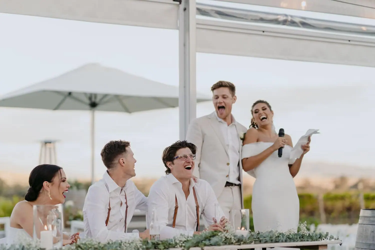 Bride holding a microphone beside the groom as their wedding party laughs.
