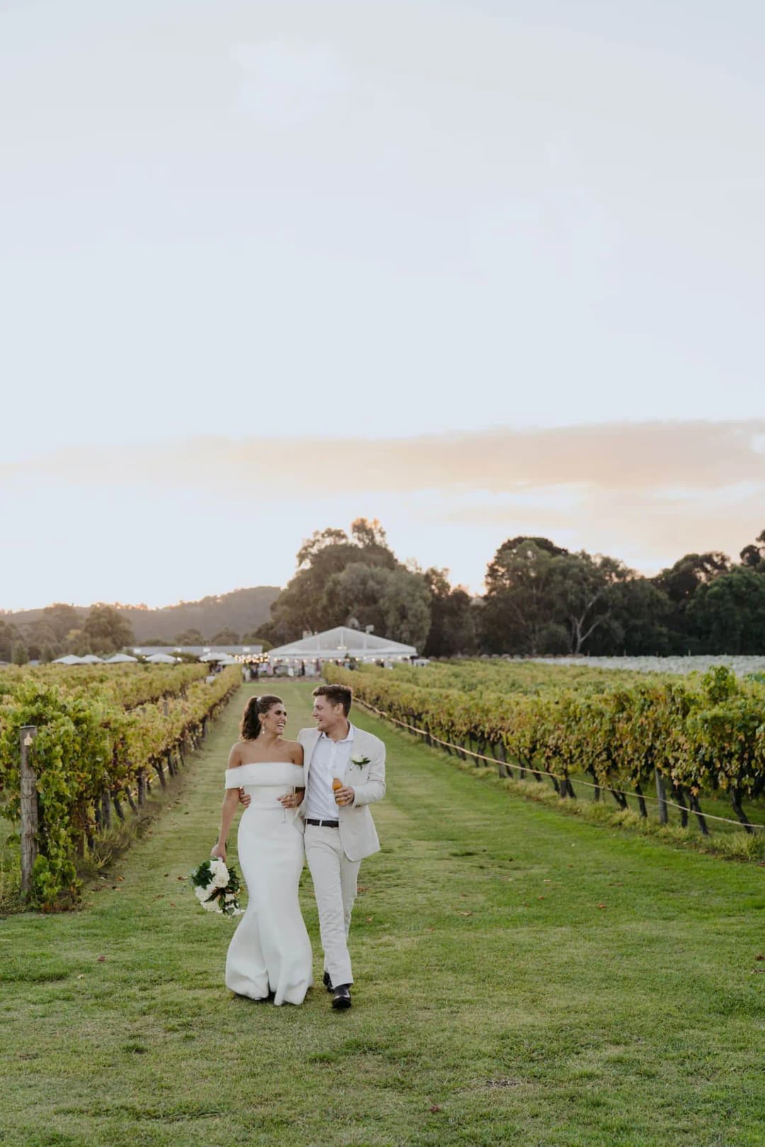 Bride and groom standing together on a grassy path between vineyard rows.