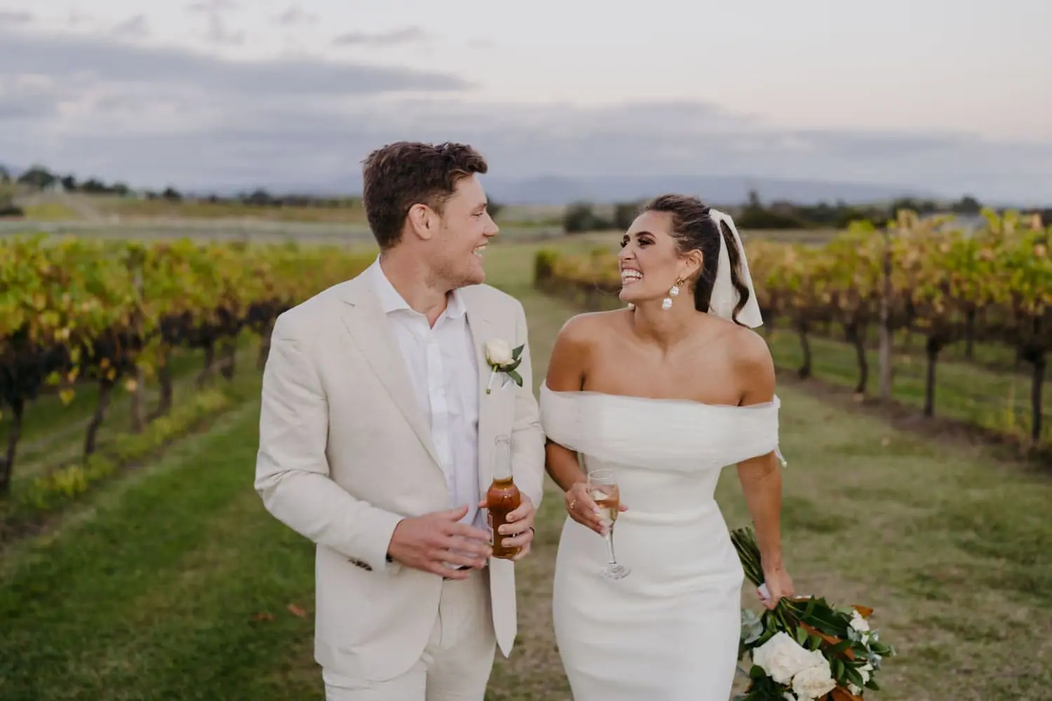 Couple smiling at each other as the groom holds two bottles beside the vineyard.