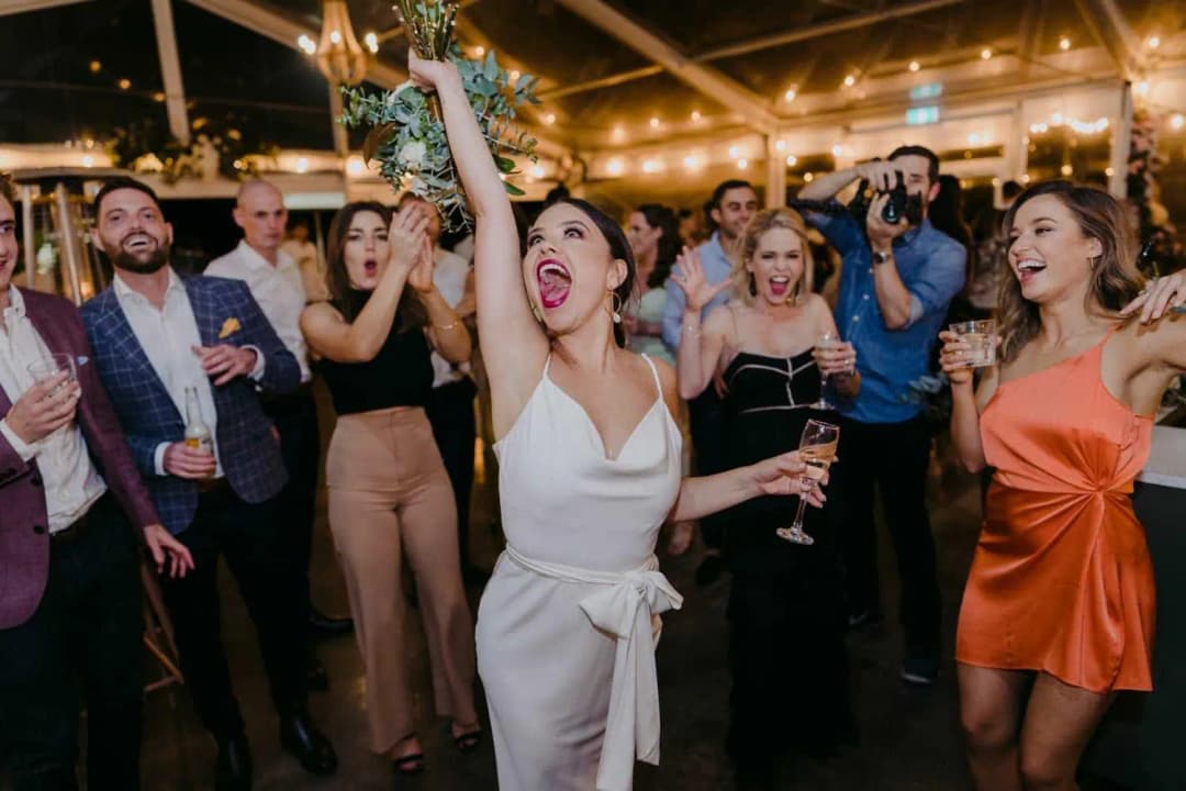 Woman raising a bouquet overhead while cheering guests surround her on the dance floor.
