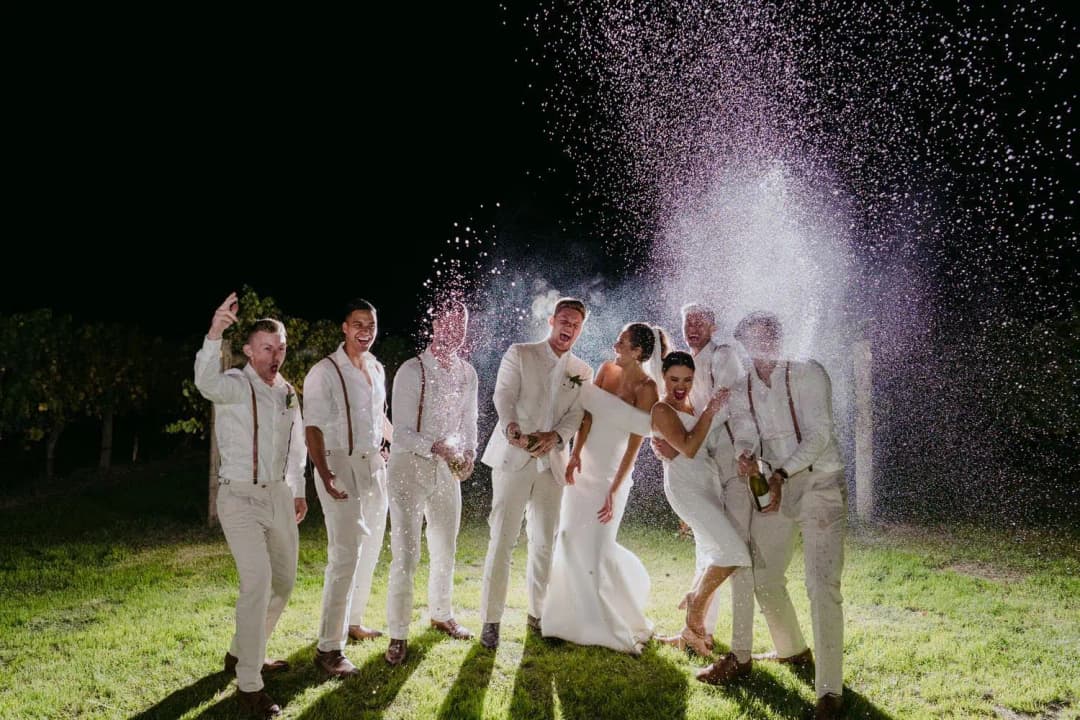 Wedding party laughing on the lawn at night as the groom and a companion spray sparkling wine.