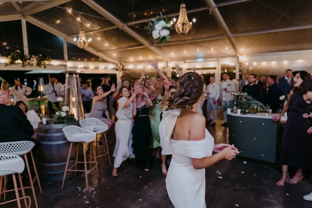 Bride holding a drink and watching guests cheer on the dance floor.