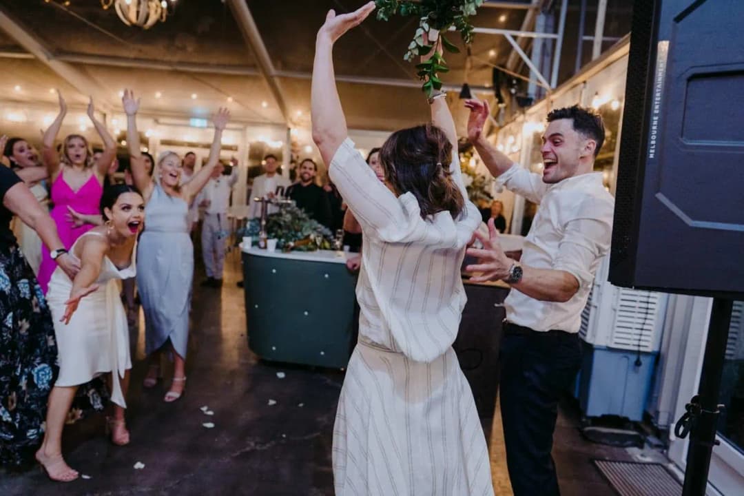 Bride raising an arm while the groom and guests cheer beside a speaker.
