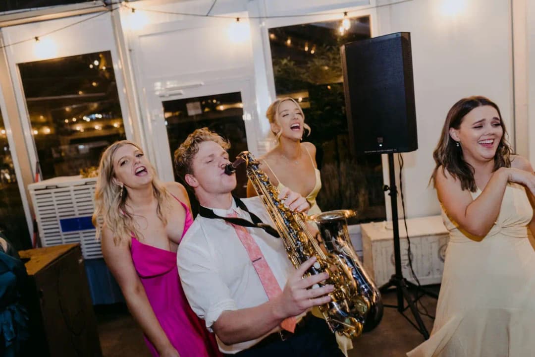 Saxophonist playing among smiling guests inside the illuminated marquee.