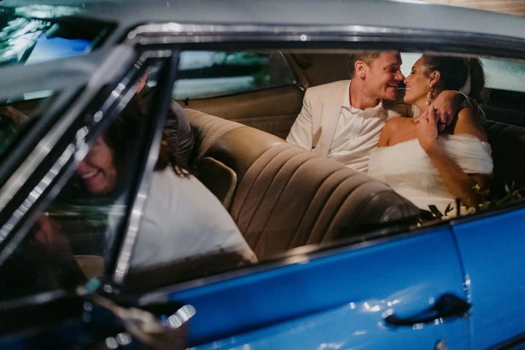 Bride and groom smiling at each other in the back seat of a blue vintage car.