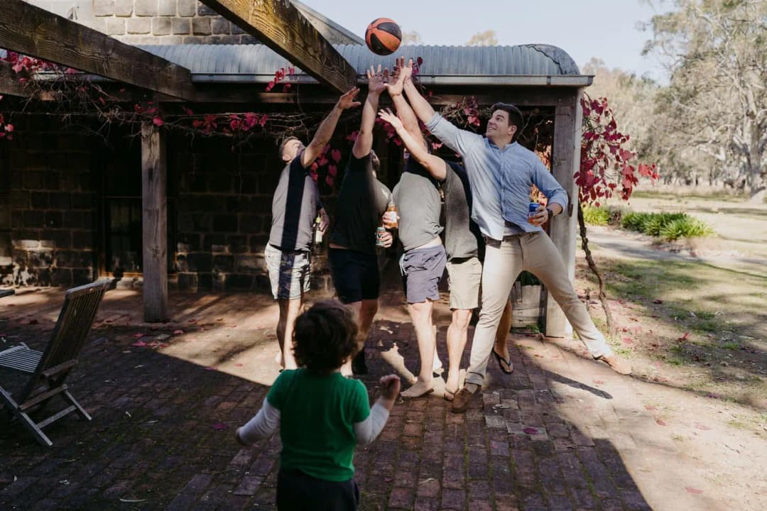 Men reaching for a basketball beneath a wooden pergola while a small child watches.