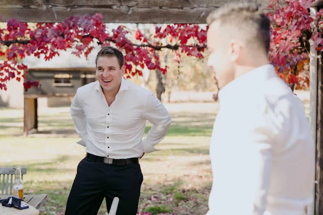 Groom fastening his white shirt beneath a pergola with red leaves.