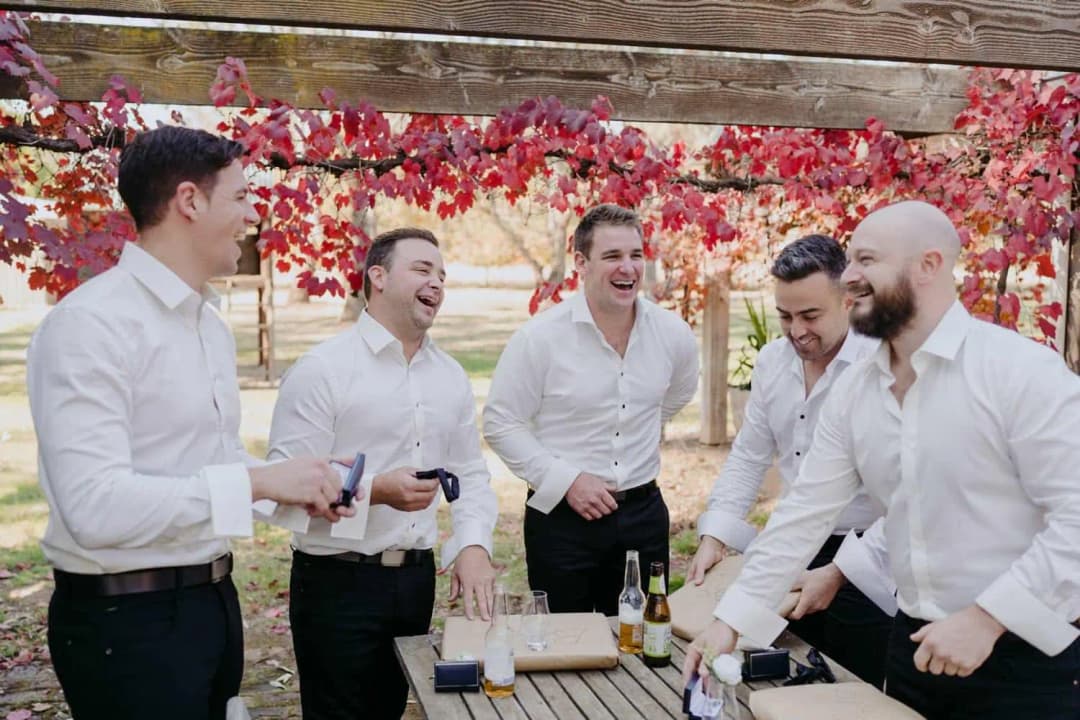 Five men laughing together in white shirts beside a table under red autumn leaves.
